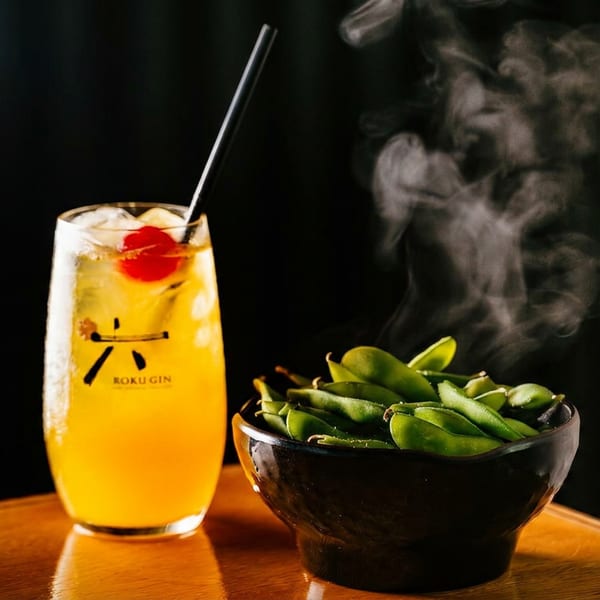 Bright orange cocktail with cherry and straw beside a steaming bowl of edamame on a wooden table.