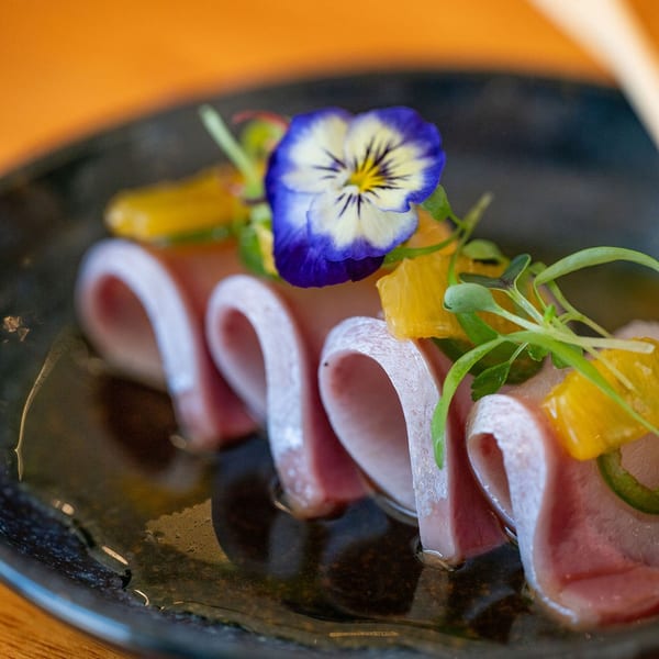 Elegant dish of rolled raw fish garnished with orange segments, microgreens, and a purple pansy on a black plate.