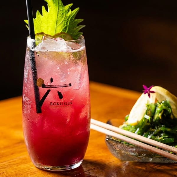 Pink iced Roku gin cocktail garnished with shiso leaves beside seaweed salad, lime wedge, and chopsticks on a wooden table.