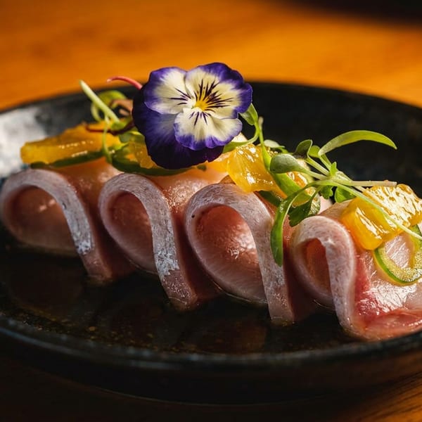 Thinly sliced raw tuna arranged on a black plate with citrus segments, greens, and an edible purple flower.