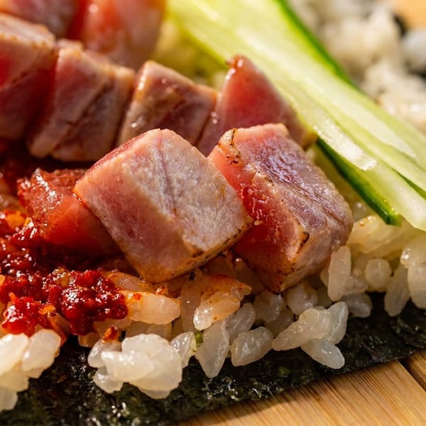 Close-up of seared tuna cubes, rice, chili sauce, and cucumber on a sheet of nori.