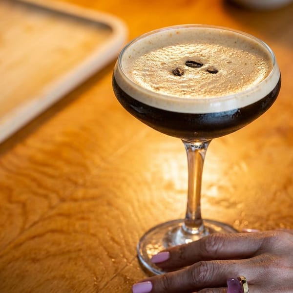 Espresso martini topped with coffee beans in a coupe glass on a warmly lit wooden restaurant table.