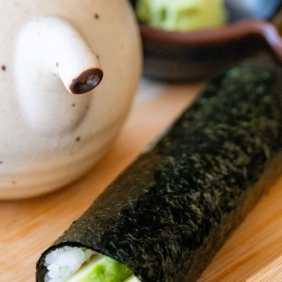 Sushi hand roll filled with white rice and avocado on a wooden tray, beside a ceramic teapot and dipping bowl.