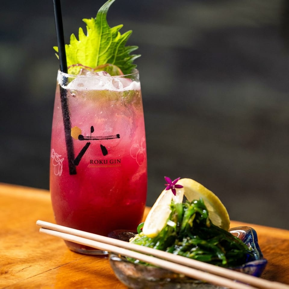 Pink Roku Gin cocktail garnished with shiso leaves beside a bowl of seaweed salad, lemon wedge, and chopsticks on a wooden table.