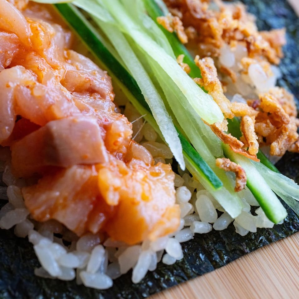 Close-up of a nori sheet topped with sushi rice, spicy salmon, cucumber strips, and crispy fried onions.
