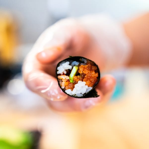Hand holding a cut sushi roll filled with white rice, orange roe or fish, and green vegetables.