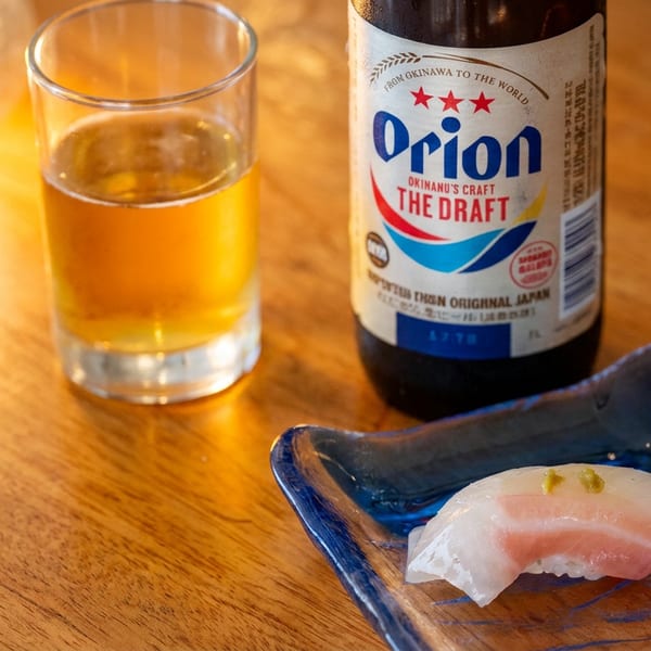 Nigiri sushi sits on a blue ceramic plate beside a glass of beer and an Orion Okinawa draft beer bottle on a wooden table.