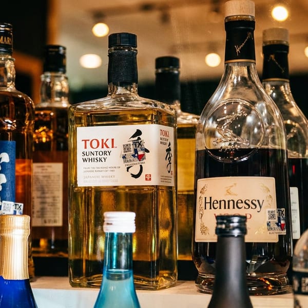 Assorted whiskey and cognac bottles, including Toki Suntory Whisky and Hennessy, displayed on a warmly lit bar shelf.
