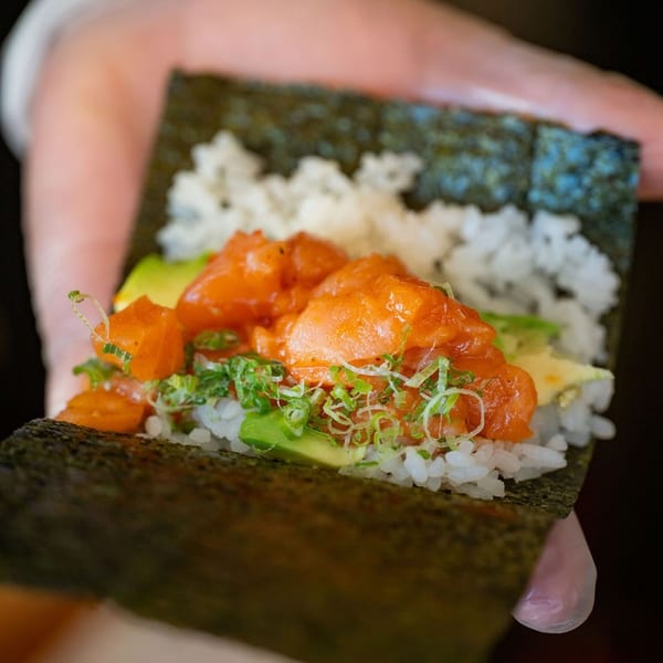 Hand-held seaweed wrap filled with sushi rice, avocado, salmon, orange sauce, and sliced green onions.