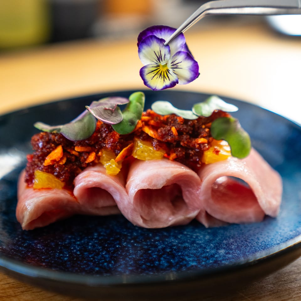 Gourmet dish of rolled pink slices topped with spiced crumble, citrus pieces, greens, and an edible purple flower on a black plate.