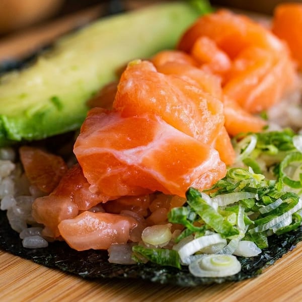 Close-up of a sushi hand roll with salmon, avocado, rice, seaweed, and sliced green onions on a wooden board.